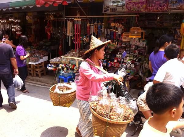 Woman in straw hat carrying baskets of snacks through a busy street market