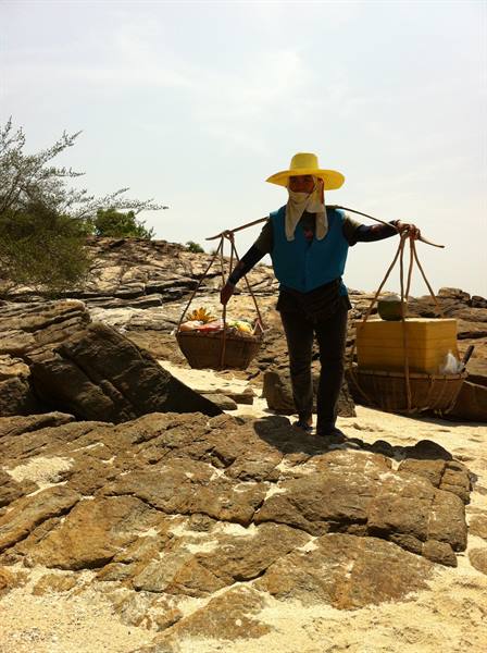 Woman in yellow hat carrying baskets on shoulder pole across rocks