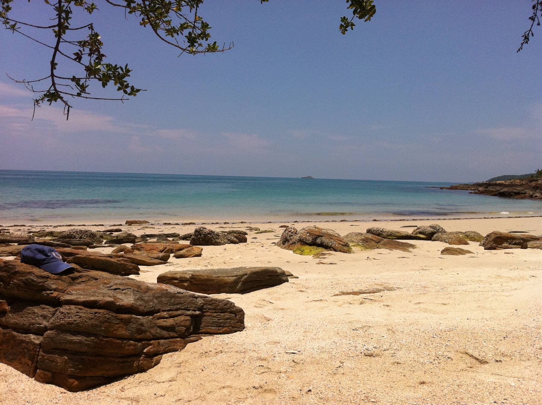 Blue cap on brown rocks, pale sand and turquoise sea under green branches