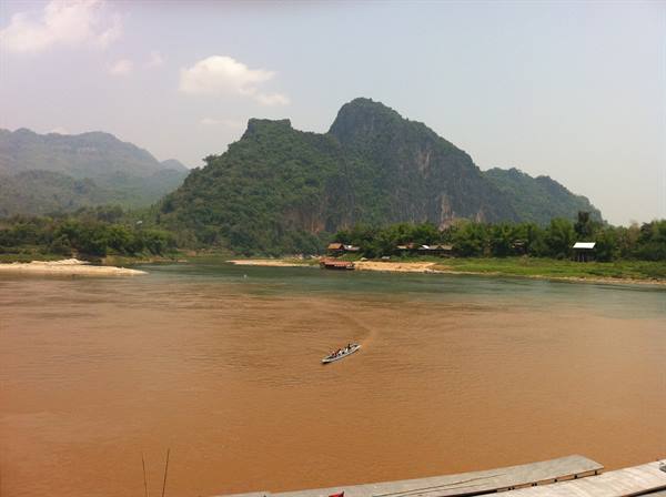 Boat crossing muddy brown river beneath green limestone hills and sandy banks
