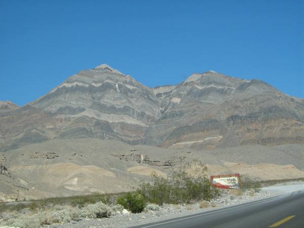 Layered rocky mountains under blue sky, desert road with sign and shrubs