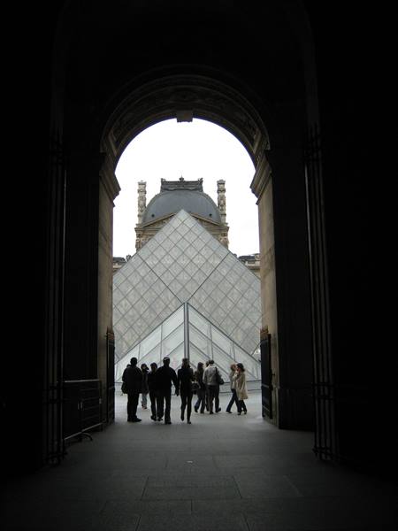 Louvre glass pyramid seen through dark archway, visitors walking outside