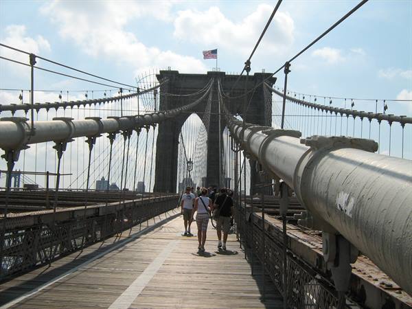 Tourists walk on Brooklyn Bridge wooden walkway among steel cables, American flag above