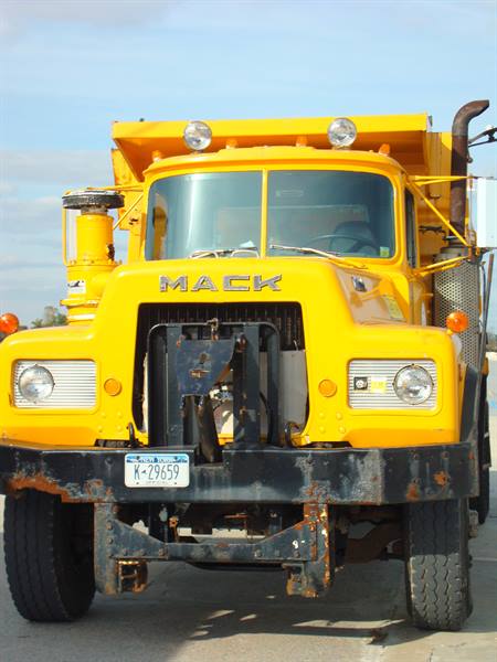 Yellow MACK dump truck with front scraper blade, headlights on, blue sky