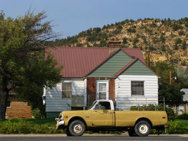 Pickup giallo d'epoca parcheggiato davanti a una casa in legno, colline rocciose alle spalle