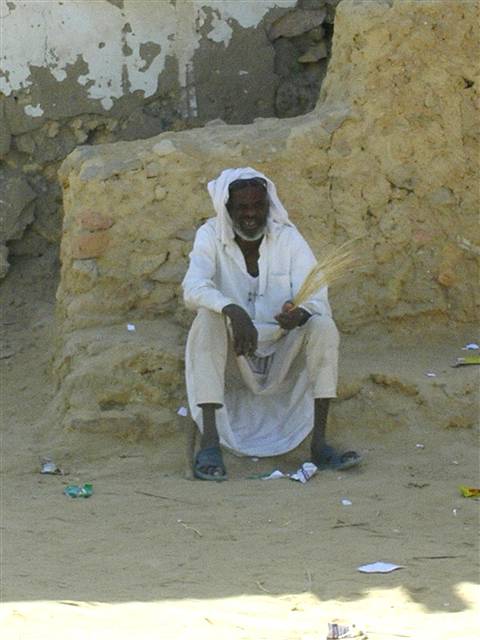Man in white robe sitting on mud steps, holding a straw bundle