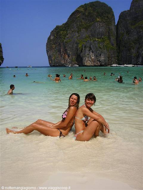 Two girls in bikinis sitting on white sand, swimmers and limestone cliffs behind