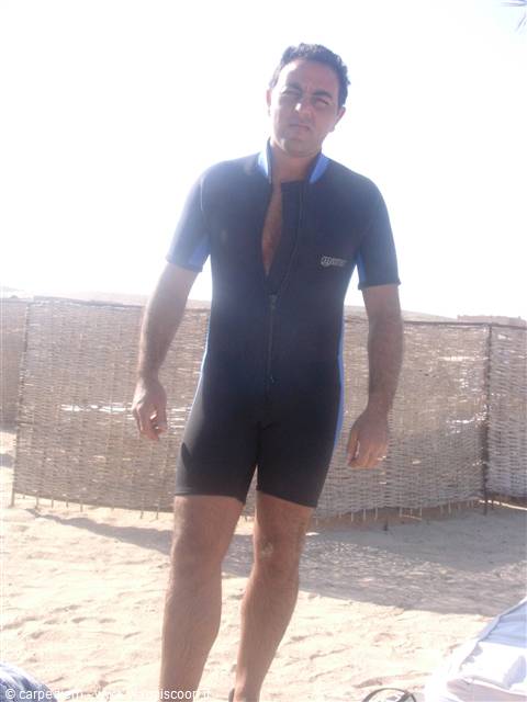 Man in black and blue short wetsuit standing on sandy beach by reed fence