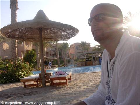 Man wearing sunglasses beside a pool with straw parasol and sun loungers
