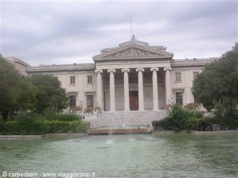 Neoclassical columned palace in front of a fountain among greenery, grey sky