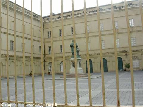 Statue in paved courtyard behind yellow bars, ochre buildings with green doors