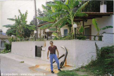 Man in t-shirt and jeans next to an iron anchor by a house