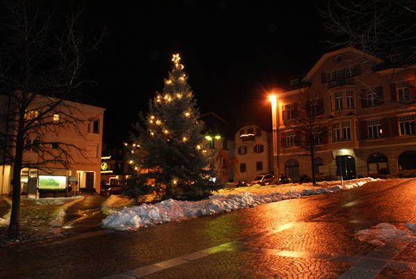 Illuminated Christmas tree on snowy cobblestone square, alpine buildings at night