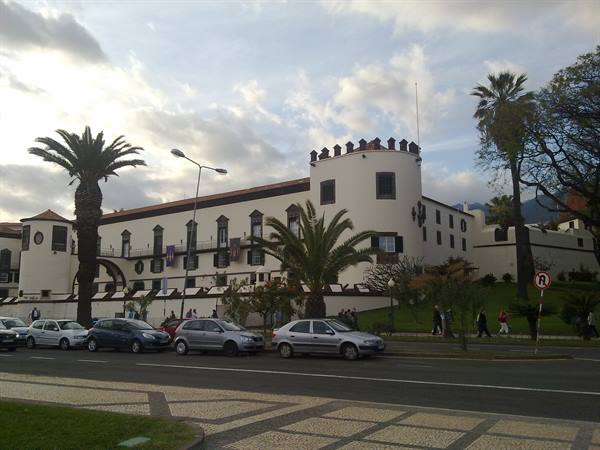 White crenellated palace building with palm trees and parked cars on the street