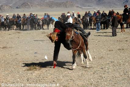 Rider leaning low on galloping bay horse, crowd of horsemen on steppe