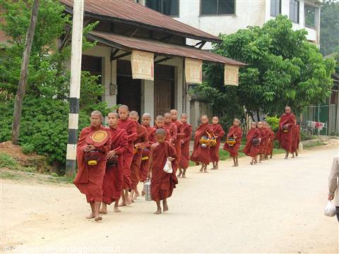Line of Buddhist monks in red robes with alms bowls walking on dirt road