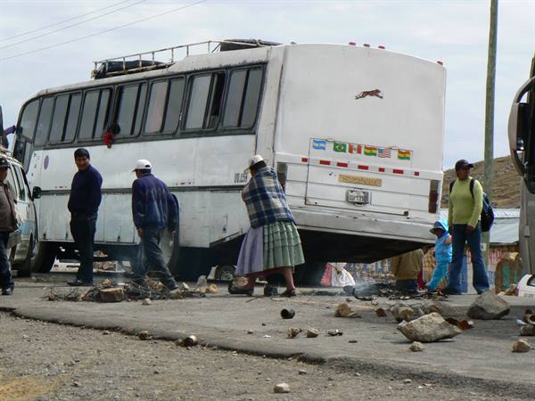 White bus stopped on dirt road, women in traditional skirts among pedestrians and rocks