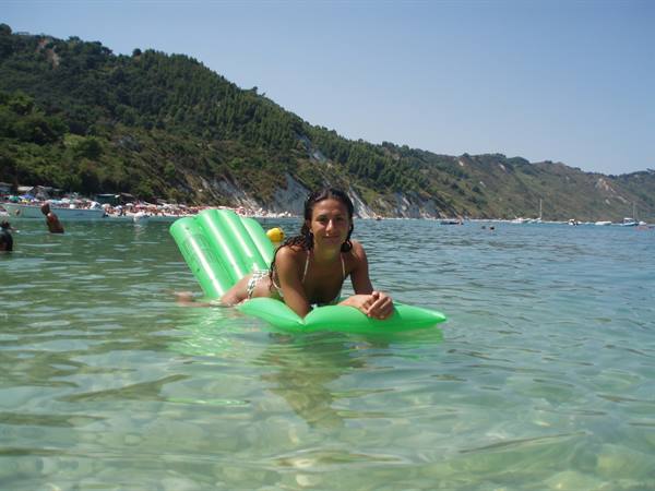 Woman in bikini on a green inflatable raft, clear water, crowded beach below green hills