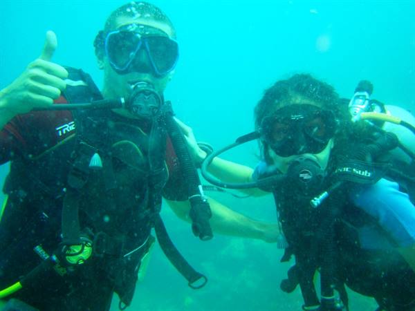 Two scuba divers with tanks and masks underwater in green water, one gives thumbs up