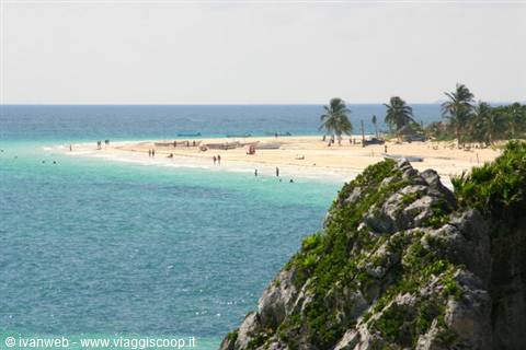 White sand beach with palm trees, swimmers in turquoise water, green rocks below
