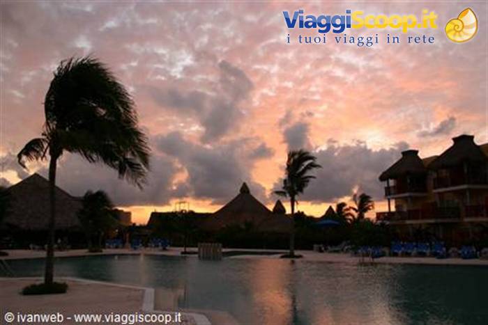 Palms and thatched roofs around the pool under pink glowing clouds