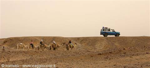 Tourists on camels cross the desert, a blue jeep watches nearby