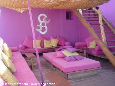 Rooftop lounge with pink sofas and yellow cushions under straw canopy, wooden stairs