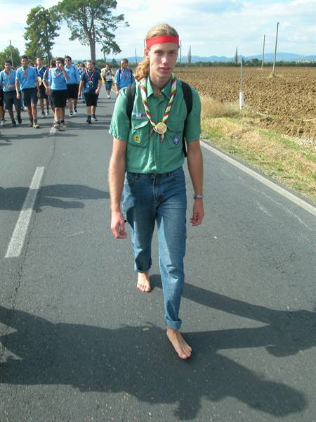 Barefoot scout with red headband and medal walking on asphalt, group behind