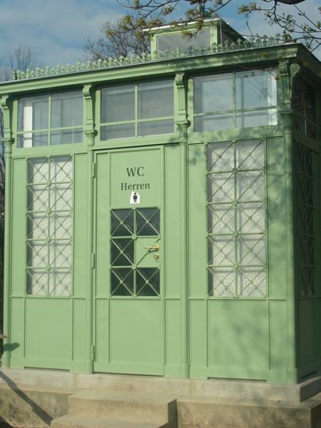 Green wooden and glass WC kiosk, Herren sign, sunlight filtering through branches