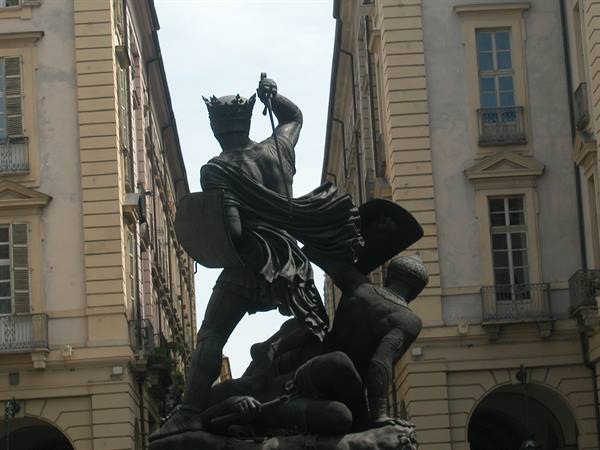 Bronze statue of crowned king raising sword over fallen enemies, between historic buildings