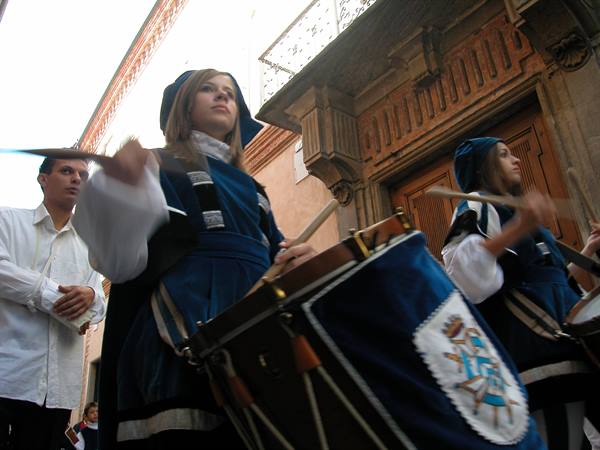 Girls in blue medieval costumes play drums in front of a stone building