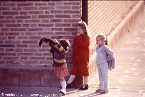 Three children playing beside a brick wall, one lifting a sweater
