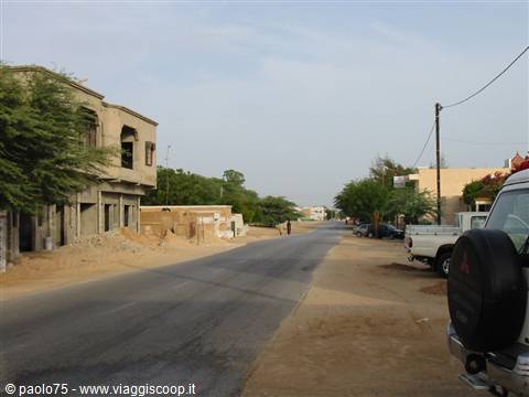 Dirt road through a village with concrete buildings, parked van, sand and trees