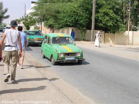 Tourists walk beside a green Renault 4 taxi on a sandy street