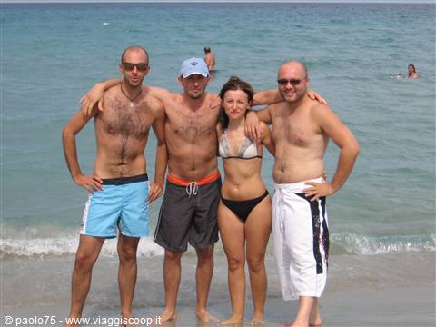 Four friends in swimwear pose arm in arm on the beach, sea and swimmers behind