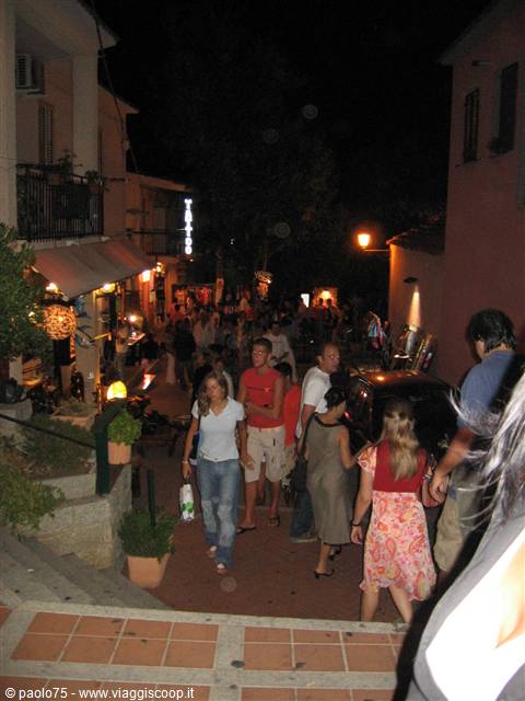 Evening strollers walk past lit-up shops on a sloping village street