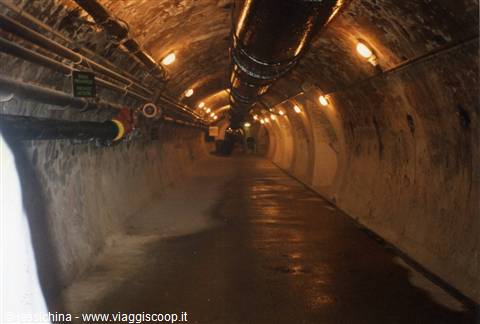 Rock-hewn tunnel with pipes and yellow lights, wet floor