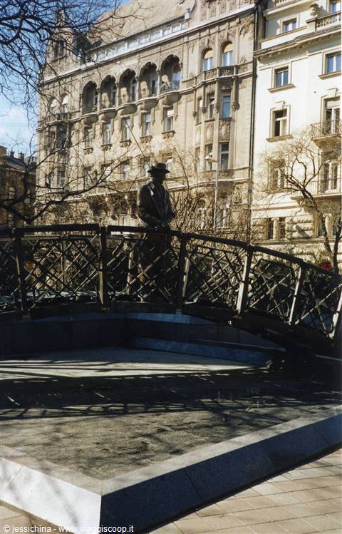 Statue in coat and hat on iron bridge, Art Nouveau buildings behind