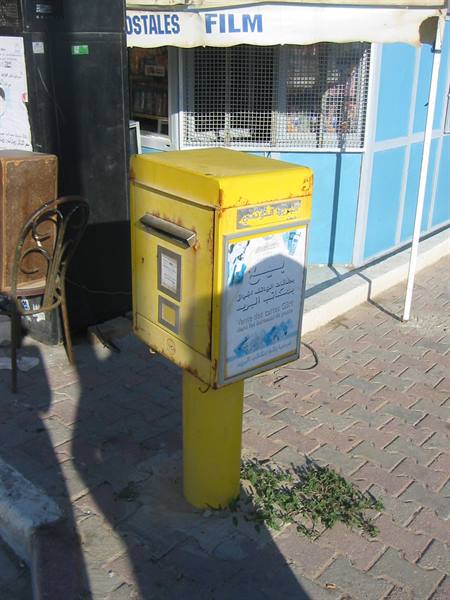 Yellow mailbox on paved sidewalk, postal kiosk in the background