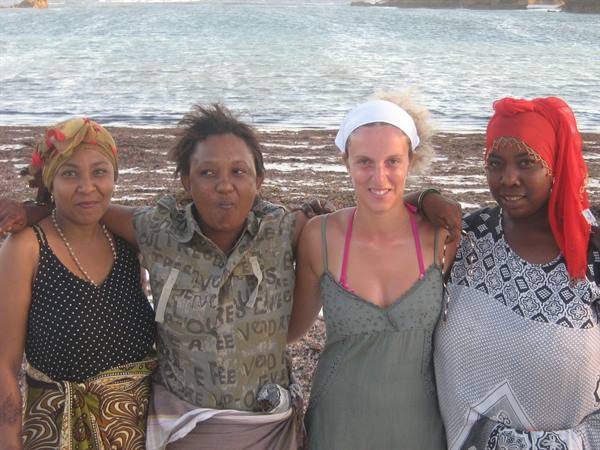 Four women arm in arm on the beach, colorful headscarves, rippling sea behind