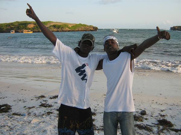 Two young men raising arms on the beach, boats and waves behind