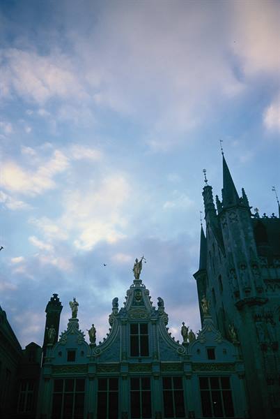 Gabled facade with golden statues and spire under clouds, Bruges