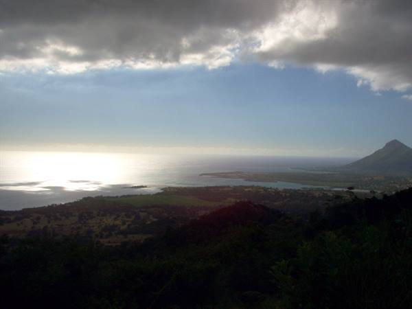 Green coastline under dark clouds, sunlight reflecting on the sea beside a mountain