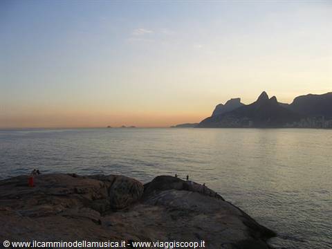 People sitting on rocks by the sea with Morro Dois Irmãos in Ipanema
