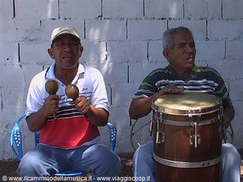 Two musicians playing maracas and a wooden drum in front of a concrete block wall
