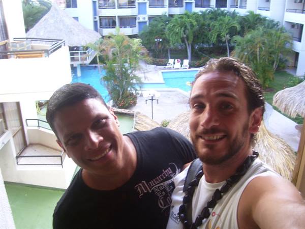 Two smiling friends on the balcony, tropical pool with palms and palapa behind