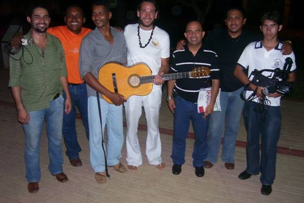 Group of smiling men at night, one holding a classical guitar, another a video camera