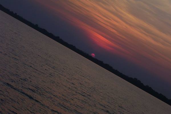 red sun sinking behind the treeline over dark rippled water