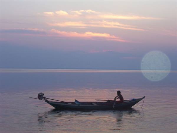 Fisherman rows a long wooden boat on calm pink-lit water at dawn