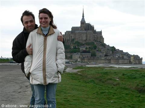 Couple hugging in white jacket in front of Mont Saint-Michel abbey, grey sky